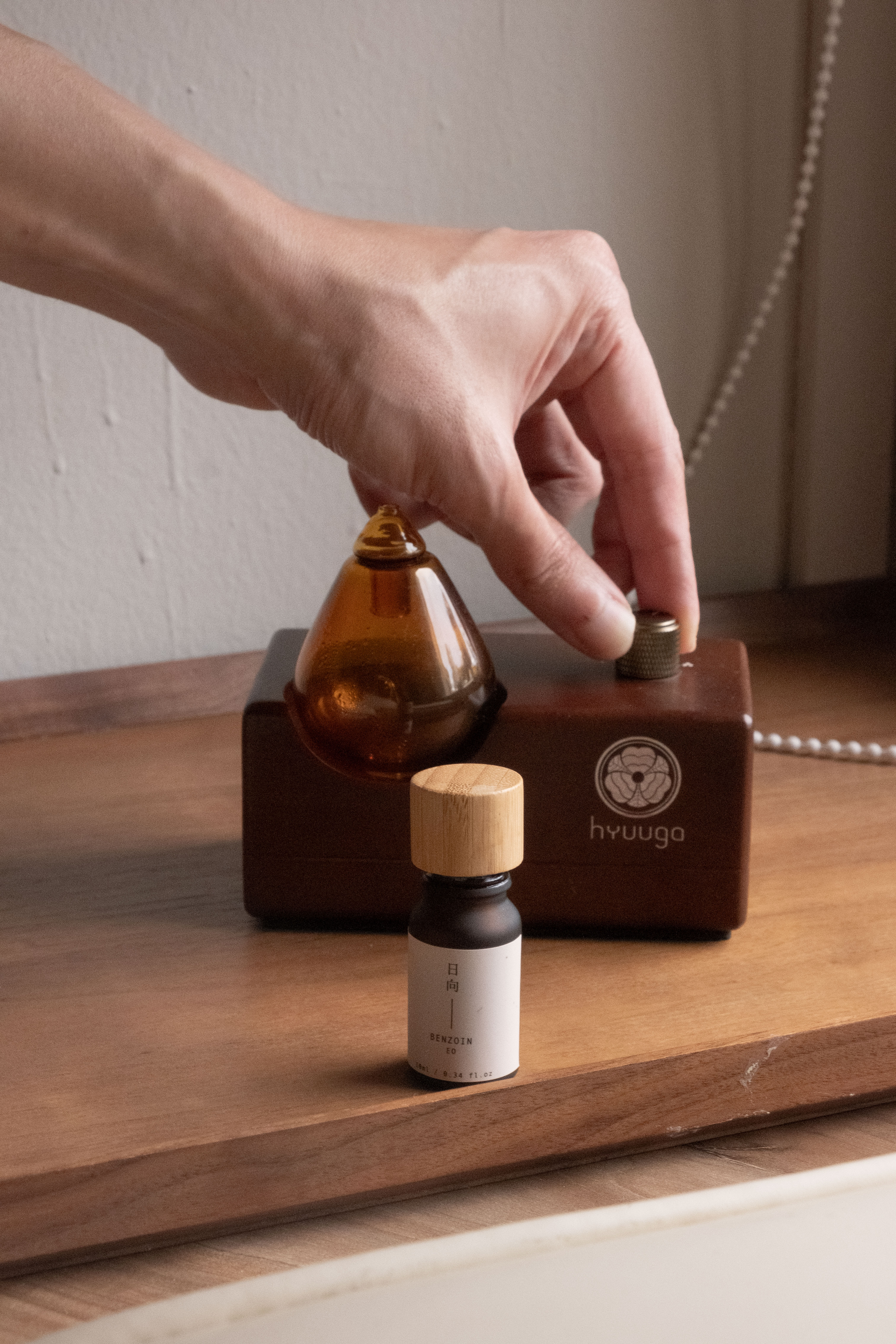 Hand twisting knob on a wooden Shinrinyoku Home diffuser, with a bottle of Benzoin Essential Oil.