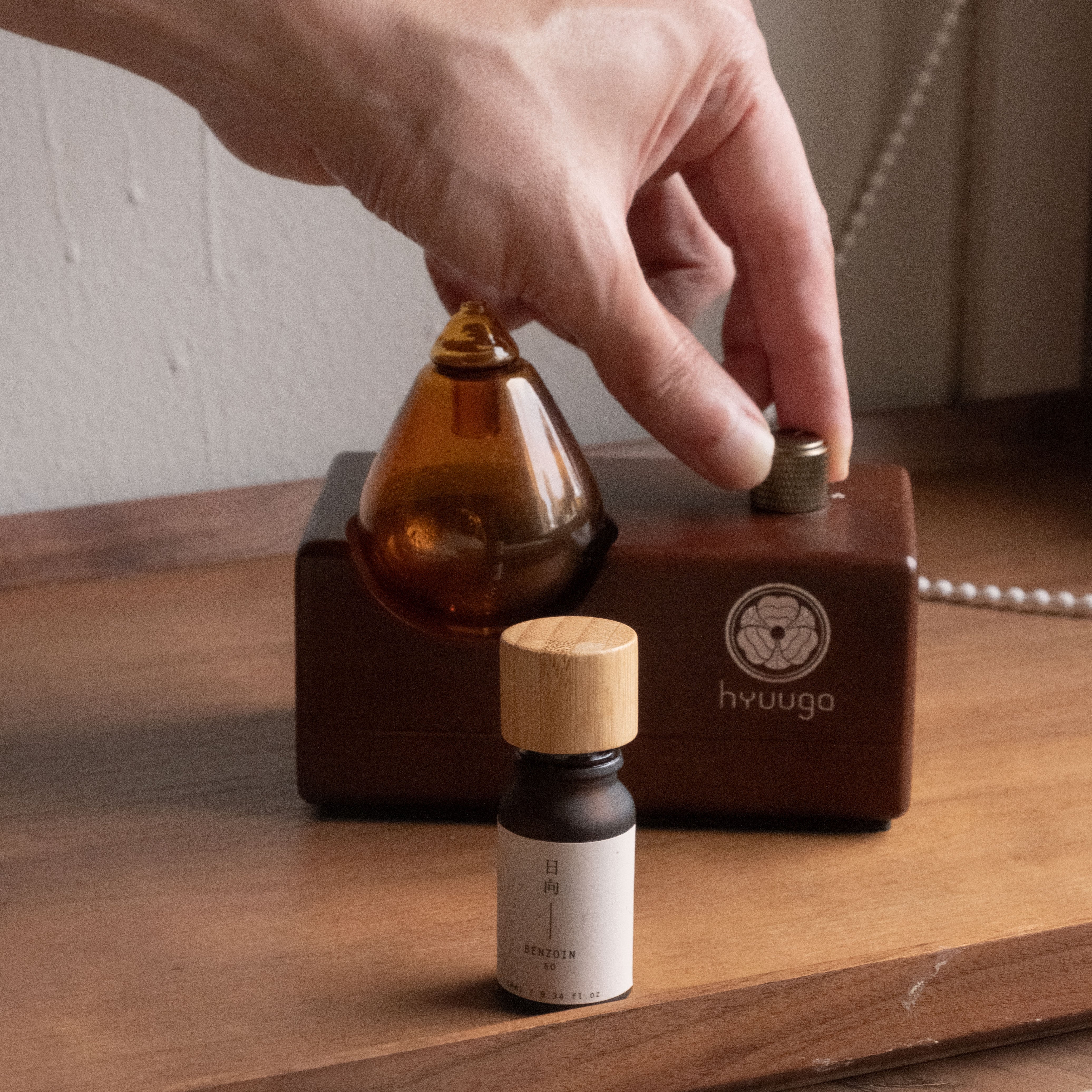 Hand twisting knob on a wooden Shinrinyoku Home diffuser, with a bottle of Benzoin Essential Oil.
